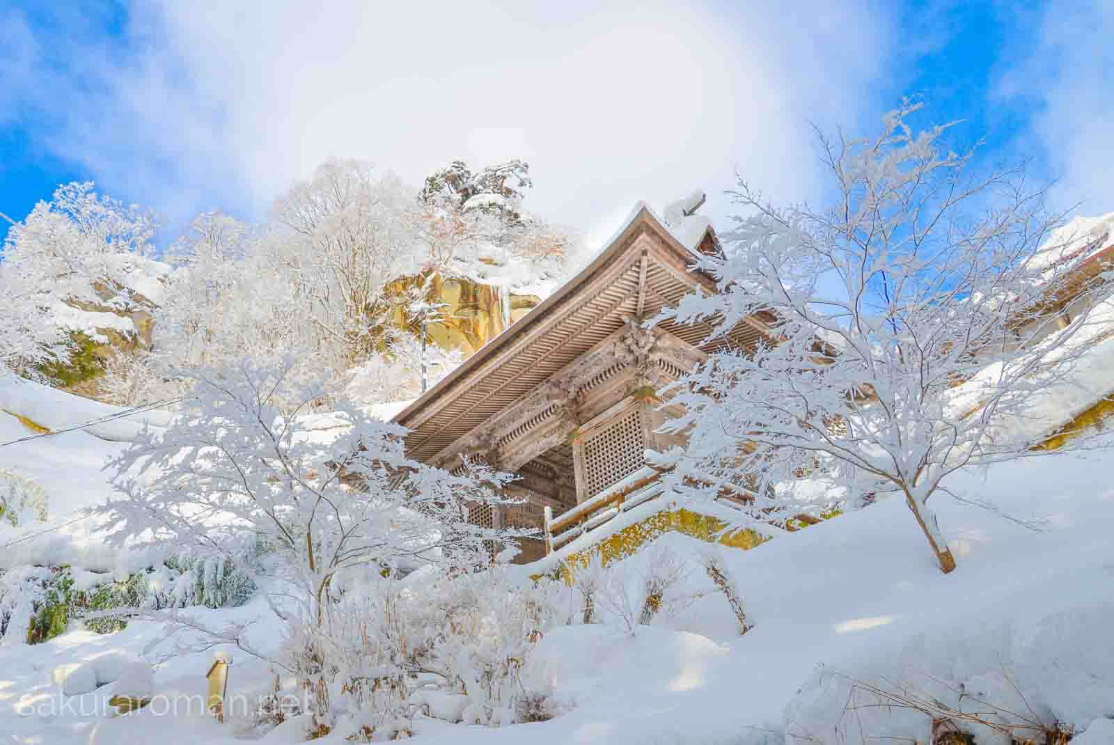 冬の山寺の雪景色に映える仁王門。青い空と白い雲、雲が続いたような雪景色の白に山門と岩肌が金色で爽やかに見える。